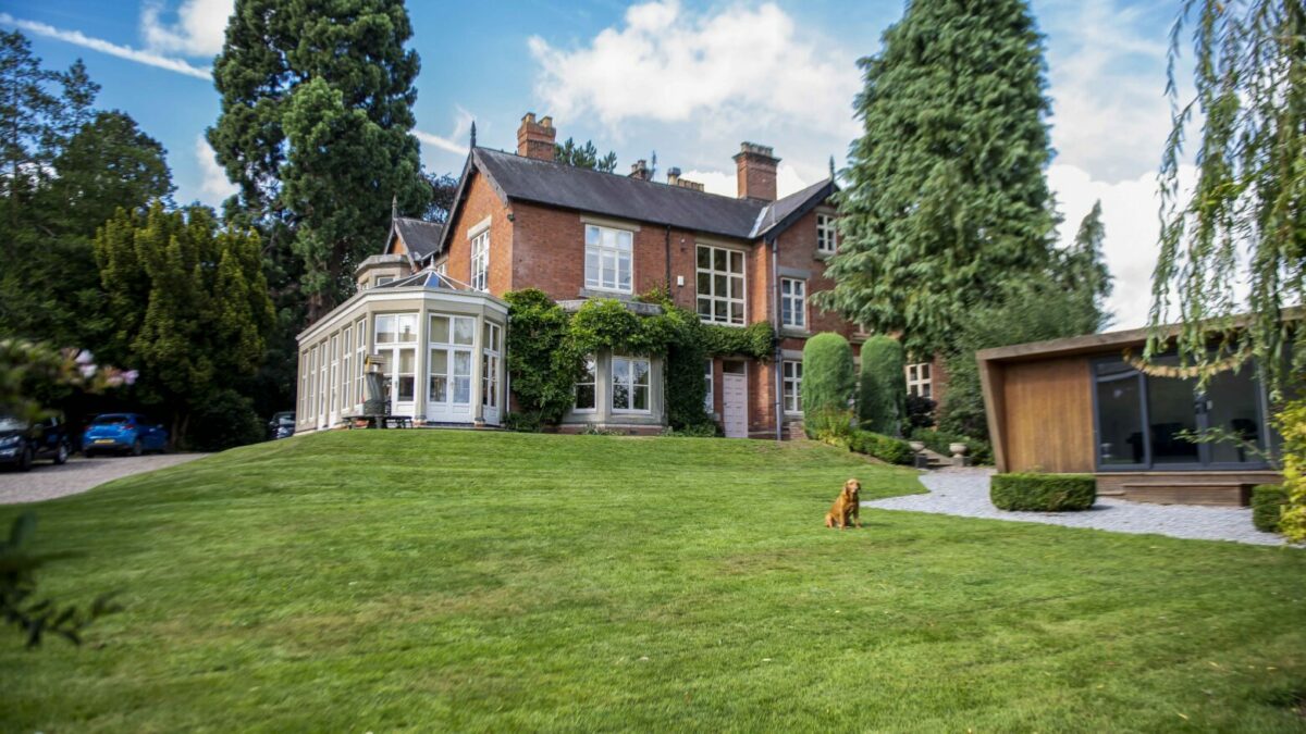 Large red-brick manor with a manicured lawn, tall trees, and a golden retriever sitting on the grass in the foreground.