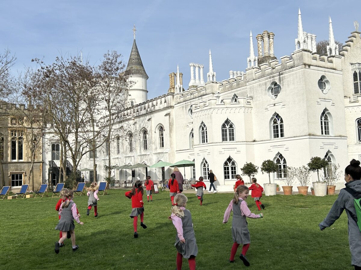 Children in red and gray uniforms run and play on a green lawn beside a white castle-like building with towers and arched windows.